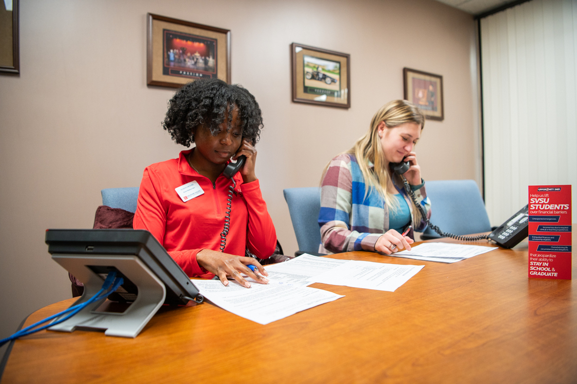 Two students sit at table, talking on black phones
